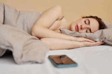 Rahat bir yatakta uzanan ve akıllı telefonuyla uyuyan genç bir kadının minimum portresi.