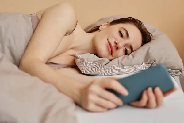 Minimal portrait of young woman lying in comfortable bed and using smartphone scrolling social media
