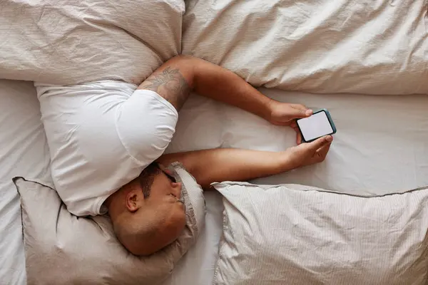 Minimal top view of black man lying in bed and holding smartphone with white screen mockup, copy space