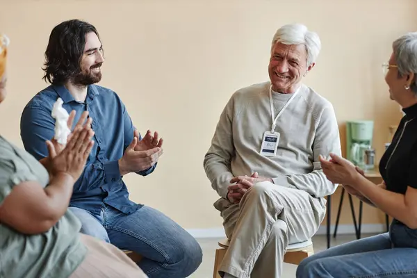 Group of smiling senior people applauding to white haired man sitting in circle at mental health support group and sharing success stories