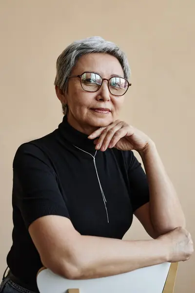 Waist up portrait of modern Asian senior woman looking at camera confidently leaning on chair