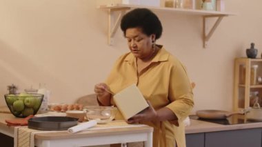 Yaşlı siyahi bir kadının, evde sıcak bej bir mutfakta yemek pişirirken cam kaseye koyduğu kuru yiyecek kutusundan biraz un eklerken orta boy fotoğrafı.