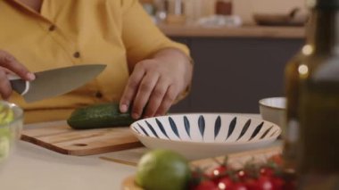 Yaşlı siyahi bir kadının yemek masasında oturup taze salatalıkları bıçakla kesmesi ve evde öğle yemeği için sağlıklı sebze salatası yapması.