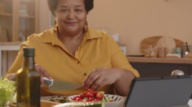 Afrika kökenli Amerikalı orta yaşlı kadının belini kaldırarak kameraya gülümserken günışığıyla aydınlanan mutfakta sağlıklı sebze salatası yapıyor.