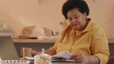 Orta yaşlı, zarif siyahi bir kadının kahvaltı sırasında mutfak masasında gazete okuduğu ve arkadaşlarıyla dijital tablette görüntülü sohbet yoluyla haberleri tartıştığı bir fotoğraf.