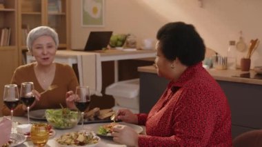 Çok ırklı iki yaşlı kadın ve erkeğin sohbet ettiği, şarap içtiği ve sıcak bir dairede yemek yediği orta boy bir fotoğraf.