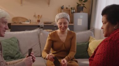 Üç çok ırklı kıdemli arkadaşın gündüz vakti güzel bir evde kahve masasında oturmuş kart oynarken eğlendikleri orta boy bir fotoğraf.