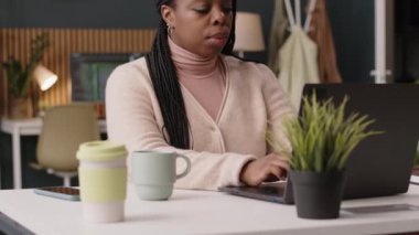 Genç Afrikalı Amerikalı bir kadının dizüstü bilgisayarının yanında otururken yazmasını, sürdürülebilir yeşil ofis alanında çalışmasını.