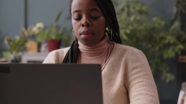 Genç Afrikalı Amerikalı kadın ofis çalışanının göğüs röntgeni. Bulanık arka planda karbondioksit azaltmak için ev bitkileriyle çağdaş iş yerinde dizüstü bilgisayar kullanıyor.
