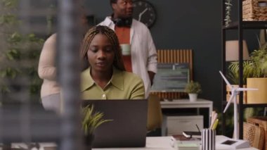 Orta boy, siyah bir kadının laptopun önünde, sürdürülebilir yeşil ofis alanında, arka planda kahve molası sırasında iş arkadaşlarıyla sohbet ederken görüntüsü.