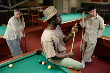 Bilardo kulübünde çok ırklı insanlarla şapka takan Afrikalı Amerikalı yetişkin bir adamın portresi.