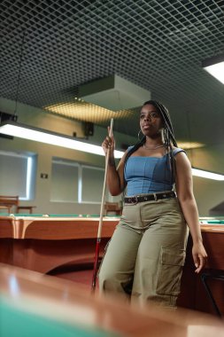 Kendine güvenen Afro-Amerikan kadının dikey düşük açılı portresi bilardo masasında duruyor ve elinde ıstaka çubuğu var.