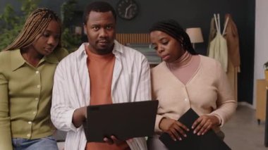 Bilgisayara bakıp kameraya poz veren ve arka planda birçok ev bitkisi olan sürdürülebilir yeşil ofiste çalışan üç kişilik siyah bir ekibin orta yavaş portresi.