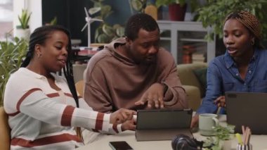 Siyah yaratıcı ekibin dijital tablet ve dizüstü bilgisayarın başında yeşil çalışma alanındaki masada oturup beyin fırtınası yaparken orta ölçekli görüntüsü.