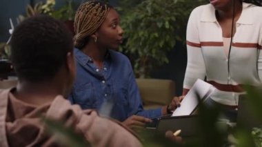 Genç Afro-Amerikan iş adamlarının modern ofiste yeşil bitkilerle yapılan gayri resmi toplantı sırasında proje detaylarını tartışırken orta ölçekli bir fotoğrafı.