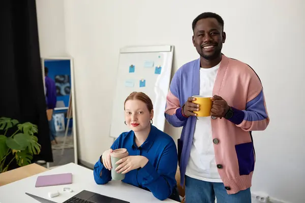 Portrait of two creative business people smiling at camera in office and holding coffee cups, vibrant colors
