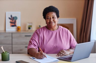 Pembe tişörtlü, gülümseyen bir kadın ev ortamında çalışırken kameraya bakıyor ve belgelere not alıyor.