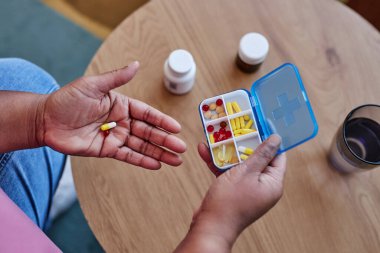 Elinde küçük kapsül ve konteynır olan yaşlı bir kadın, iki plastik şişe ve bir bardak suyla ahşap masanın üzerine haplar koyuyor.