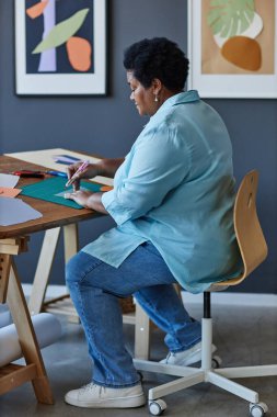 Olgun Afro-Amerikalı kadın tasarımcı veya sanatçı çeşitli malzemelerle birlikte sandalye sıra otururken yeni kağıt uygulaması üzerinde çalışıyor