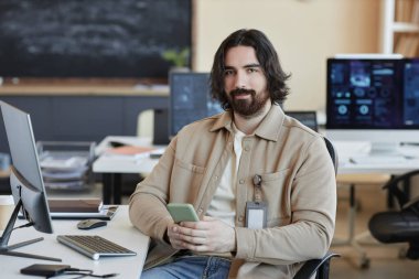 İş yerinde oturup yeni yazılımlar üzerinde çalışırken akıllı telefonlu mesajlar atan ve kameraya bakan genç sakallı bilişim mühendisi