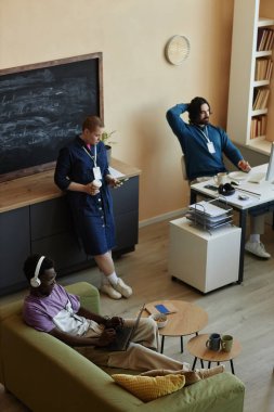 Üç genç kültürler arası yönetici molada mobil cihazlar kullanırken bir tanesi kanepede oturup dizüstü bilgisayar ekranında online video izliyor.