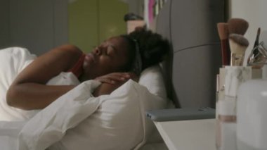 Genç uyuyan siyahi kadın sabahın erken saatlerinde yatakta uyanır, komodindeki akıllı telefona uzanır, zamana bakar ve uykuya dalardı.