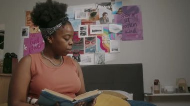 Orta halli, zeki siyahi bir kadının üniversitede ders kitaplarındaki sayfalara bakarken rahat yatak odasında, duvarında parlak posterlerle ev ödevi yaparken çekilmiş bir fotoğrafı.