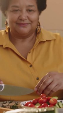 Dikey eğim, orta yaşlı Afro-Amerikan bir kadının parlak güneş ışığında sağlıklı sebze salatası yaparken kameraya gülümseyen portresi.