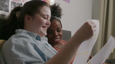 İki etnik çeşitliliğe sahip kız öğrencinin kolej ödevlerini birlikte yapmaları, birlikte yaşadıkları evde yataklarında kağıt işleriyle ilgilenmeleri ve konuşmaları.