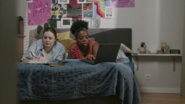 İki farklı etnik kökenli genç kız öğrenci yataklarında rahat bir yatak odasında sırt üstü uzanmış duvarlarında parlak posterlerle birlikte sınavlara hazırlanırken dizüstü bilgisayar kullanıyorlar.