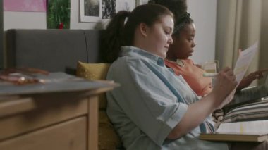 Evde araştırma projesine hazırlanan iki çok ırklı kız öğrencinin, rahat bir yatak odasında oturup, çalışma kağıtlarına yazı yazıp tartışmalarının orta boy fotoğrafı.