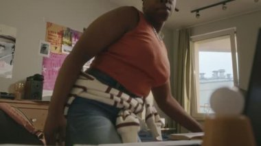 Orta yaşlı, Afro-Amerikan üniversiteli bir kız öğrencinin parlak kampüs odasında masasında oturup ödev yapmaya hazırlandığı bir fotoğraf.