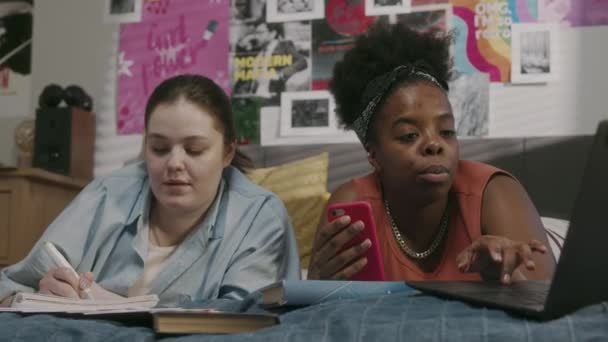 两个多民族的女朋友学生躺在明亮的现代公寓里舒适的床上 为大学做家务 聊着天 — 图库视频影像