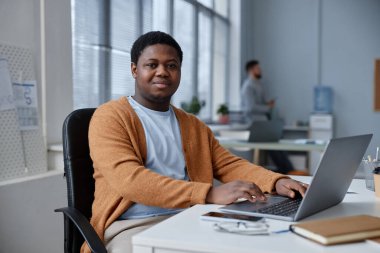 Gündelik kıyafetli genç bir adam iş yerinde dizüstü bilgisayarın önünde oturmuş internetteki bilgileri analiz ederken kameraya bakıyor.
