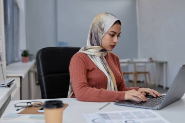 Genç, ciddi Müslüman kadın başörtüsü uzmanı yeni proje üzerinde çalışıyor ya da dizüstü bilgisayar klavyesinde yazarken çevrimiçi verileri analiz ediyor