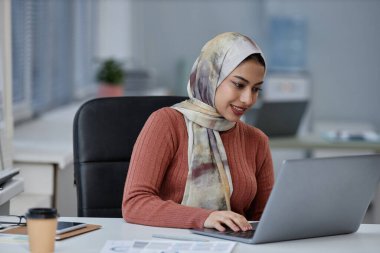 Genç, gülümseyen, başörtülü, iş yerinde dizüstü bilgisayarın önünde oturan ve ekrandaki internet bilgilerine bakan Müslüman iş kadını.