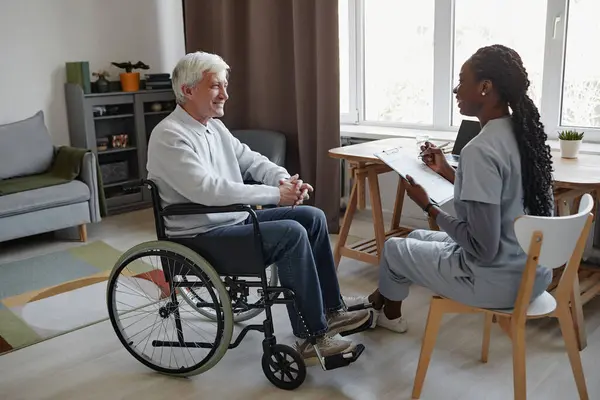 stock image Full length side view portrait of white haired senior man with disability talking to smiling nurse in retirement home, copy space