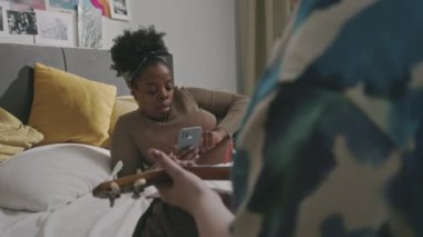 Hafta sonları evde takılırken yatakta yatan iki farklı genç bayan arkadaşın omzunun üstünden. Beyaz kız arkadaşı ukulele çalarken Afrikalı Amerikalı kız akıllı telefondan kayıyor.