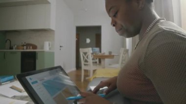 Genç Afrikalı Amerikalı bir kadının modern apartmandaki koltukta otururken bilgisayarını kullanırken, çalışırken ya da uzaktan çalışırken görüntüsünü kaldır.