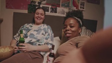 Akşamları yatakta video blogu çeken, bira içen ve yatıya kalma sırasında sohbet eden iki kaygısız kadın arkadaşın el kamerasındaki görüntüsü.