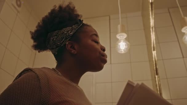 在昏暗的浴室里 年轻的非洲裔美国女人在用鞭子卷曲时 正在用纸修改学习材料 准备在大学的镜子前参加考试 拍下了一张低角度的照片 — 图库视频影像