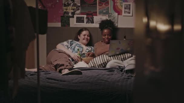 两个不同种族的年轻女性好友在床上躺在舒适的卧室里 一边喝着苹果酒 一边吃零食 晚上还在笔记本电脑上看电影 — 图库视频影像