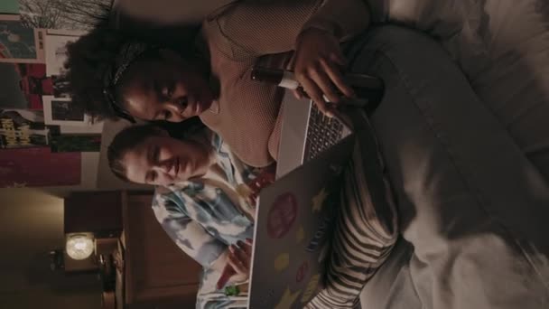 两名年轻的不同女性在晚上轻松地躺在床上喝啤酒和聊天 同时在笔记本电脑上观看系列节目的垂直镜头 — 图库视频影像