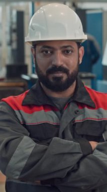 Sakallı Orta Doğu sanayi işçisinin dikey beli ve elleri bağlı kameraya poz veren iş elbisesi ve sert şapkalı portresi.