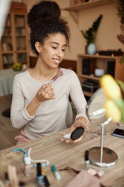Gülümseyen, kıvırcık saçlı, pencerede makyaj yapan ve aynaya bakan bir kadının dikey portresi.