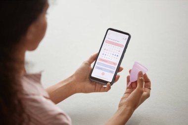 Omzunun üstünden, ekranda takvim uygulaması ve silikon adet bardağı olan akıllı telefonu tutan genç bir kadın.