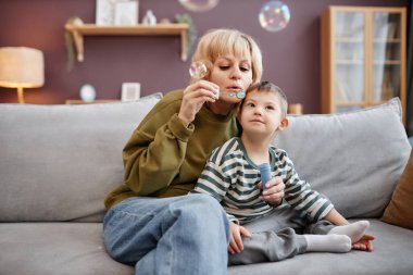 Evde kanepede otururken baloncuk üfleyen anne ve çocuk portresi.