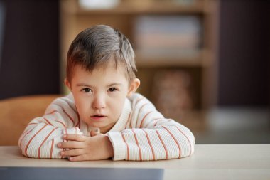 Okul masasında otururken kameraya bakan Down sendromlu ciddi bir çocuğun portresi.