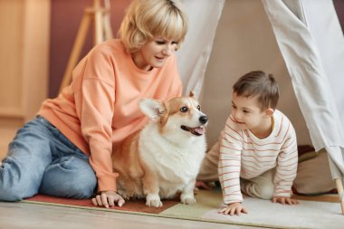 Down sendromlu küçük çocuk evde anne ve köpekle oyun oynuyor.