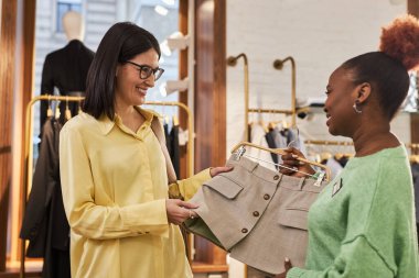 Gülümseyen kadın portresi, mağaza asistanıyla butikte kıyafetlere bakıyor.